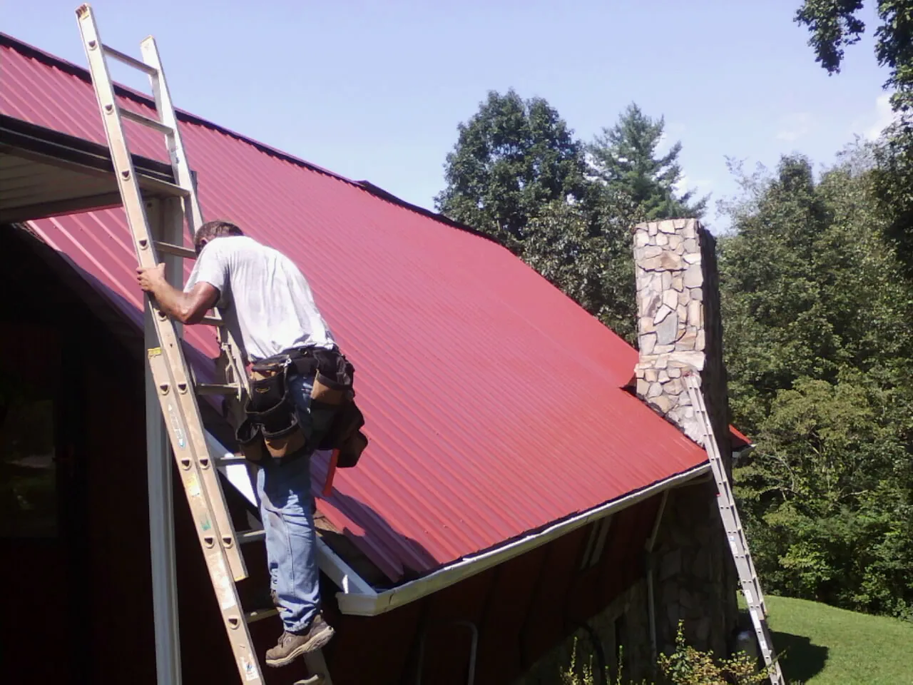 Roofer on a ladder beside a red metal roof and stone chimney — Soffit & Fascia Repair by Roofing Brothers in Waelder